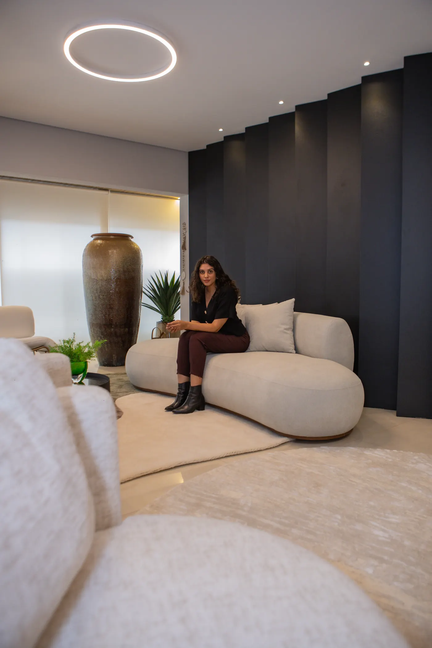 Talyssa Jade sentada no sofá curvo de uma sala de estar com painel ripado escuro e luminária circular no teto